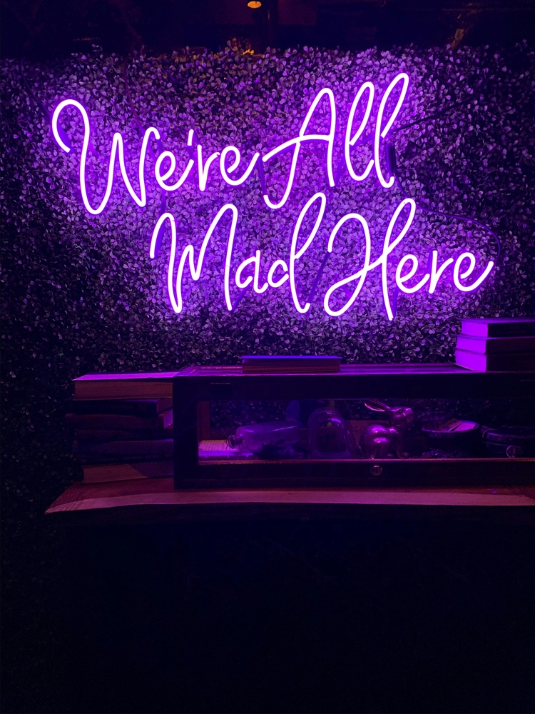 A neon sign reading 'We're All Mad Here' glowing against a wall of purple leaves